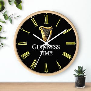 Puede incluir: Un reloj de pared negro y dorado con n&uacute;meros romanos y el texto "GUINNESS TIME" y un logotipo de arpa dorado.