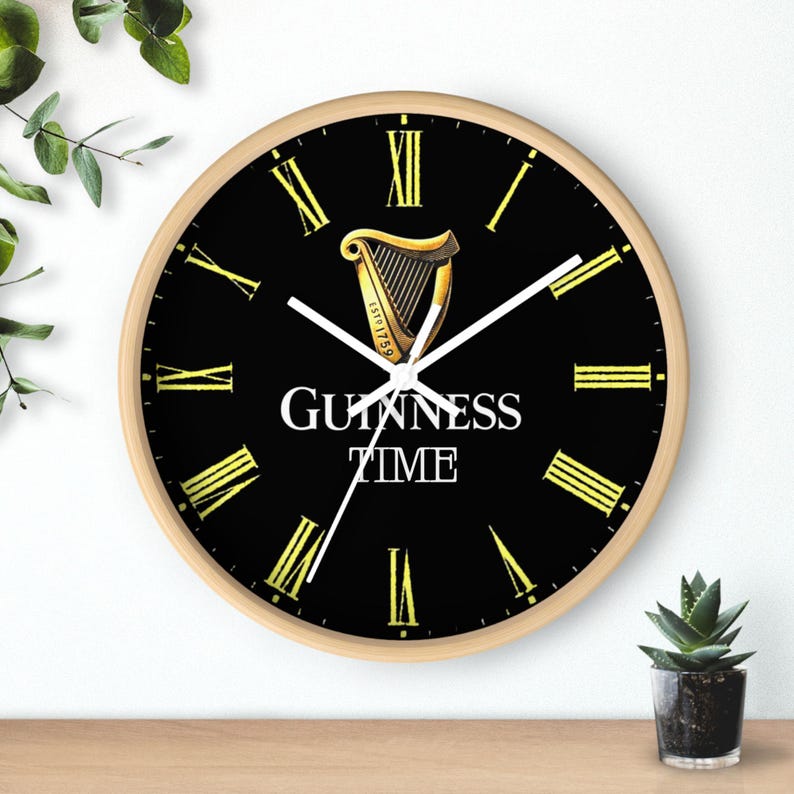 Puede incluir: Un reloj de pared negro y dorado con n&uacute;meros romanos y el texto "GUINNESS TIME" y un logotipo de arpa dorado.