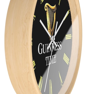 Puede incluir: Un reloj con marco de madera, esfera negra y marcadores de hora con n&uacute;meros romanos blancos. La esfera del reloj presenta un logotipo de arpa Guinness dorado con el texto "ESTD 1778" y "GUINNESS TIME".