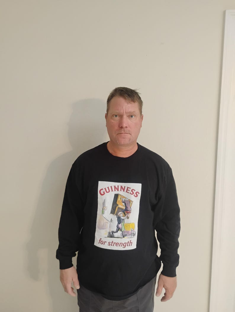 May include: Black long-sleeve shirt with a Guinness graphic. The graphic features the Guinness logo and the words "for strength" with an illustration of a man carrying a box. The shirt is a casual style.