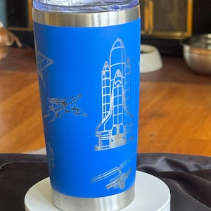 May include: A blue insulated tumbler with a silver lid and base. The tumbler features etched designs of a space shuttle, airplanes, and other space-themed elements. The tumbler is displayed on a white circular stand.