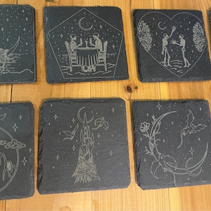 Puede incluir: Un conjunto de ocho posavasos cuadrados de pizarra con diseños grabados. Los diseños incluyen esqueletos, lunas, dragones y un castillo. Los posavasos son de color gris oscuro y están sobre una superficie de madera.