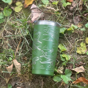 May include: A green travel tumbler with a silver rim and lid. The tumbler features an etched design of various airplanes and clouds. The tumbler is cylindrical and appears to be made of stainless steel.