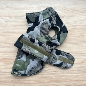Può includere: Gilet per cani con motivo mimetico nei toni del verde, grigio e nero. Presenta un'apertura per il collo e chiusure in velcro regolabili. Il nome "MESSI" è visualizzato su una piccola toppa verde, con il marchio "petico" sotto.