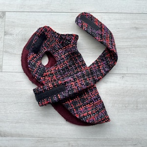 May include: A dog coat made of a woven fabric in black, purple, and red. The coat has a hole for the neck and a strap with a hook-and-loop fastener. The inside of the coat is a solid burgundy color.