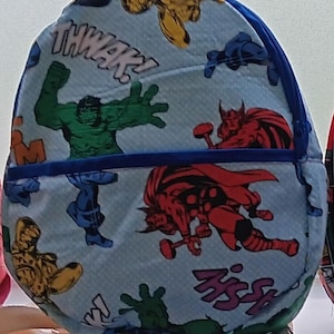 Könnte beinhalten: Ein blauer und weißer Rucksack mit Reißverschluss. Der Rucksack zeigt verschiedene Superheldenfiguren, darunter Hulk, Thor und Iron Man. Der Text "THWAK!" und "HISS!" ist auf dem Rucksack aufgedruckt.