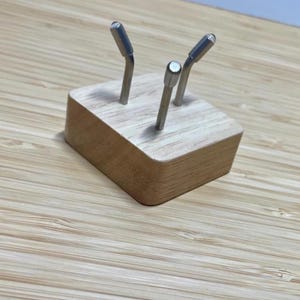 May include: A small, light-coloured wooden block with three metal rods inserted. The rods have a silver finish and are bent at the top. The block is rectangular with rounded edges. The background is a light-coloured wooden surface.