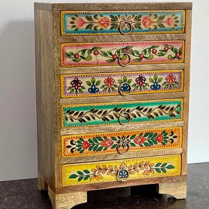 May include: A wooden six-drawer chest with hand-painted floral designs in various colours. Each drawer front features a unique pattern with a brass ring pull. The chest is made of light-coloured wood and stands on small feet.