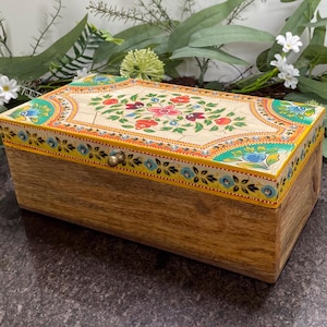 May include: A rectangular wooden box with a decorative lid. The lid has a cream background with a floral design in red, pink, and blue. The box has a brass clasp and is surrounded by greenery.