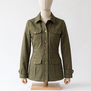 May include: An olive green utility jacket featuring a collared neckline, a front button-down closure with metallic buttons, two chest pockets with flaps, two lower pockets with flaps, and adjustable buttoned straps at the sleeve cuffs.
