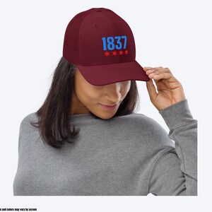 May include: A maroon baseball cap with a blue embroidered "1837" and red stars. The cap is worn by a person with long brown hair.