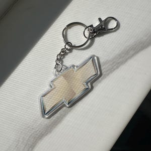 May include: A silver keyring featuring a clear Chevrolet logo charm. The charm is a light beige colour with a silver outline. The keyring has a split ring and a lobster clasp for attaching to keys or bags.