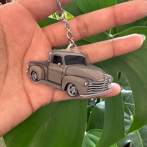 May include: A gray truck keychain with a silver key ring and chain. The truck is a classic pickup design with detailed features like the grill, headlights, and wheels. The keychain is held against a background of green leaves.