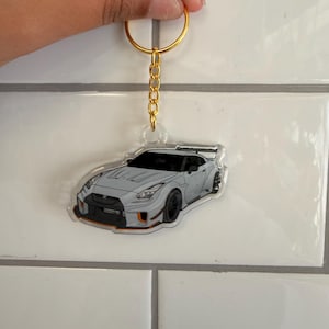 May include: A car-shaped keychain featuring a gray sports car with black wheels and orange accents. The keychain has a gold-colored chain and ring.