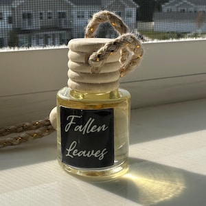 May include: A clear glass bottle with a wooden top and a woven rope handle. The bottle contains a yellow liquid and has a black label with the words "Fallen Leaves" in white script. The bottle is sitting on a white surface.