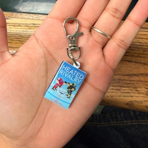 Puede incluir: Llavero de libro en miniatura con la portada de "Heated Rivalry". El libro tiene una cubierta azul con el título en blanco y rojo, y el nombre del autor. El llavero incluye un cierre plateado.