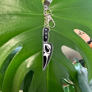 May include: A black and white knife-shaped keychain with a silver clasp. The keychain features a stylized design on the blade. The background is a large, green leaf.