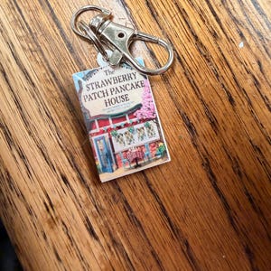 Könnte beinhalten: Ein Miniatur-Schlüsselanhänger mit einem kleinen Buch. Das Buch trägt den Text "The Strawberry Patch Pancake House" und eine farbenfrohe Illustration eines Restaurants. Der Schlüsselanhänger hat einen silberfarbenen Verschluss.