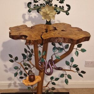 May include: A wooden table top with a tree trunk base and metal branches. The branches are decorated with green leaves and a purple snake. A yellow flower with a gold center is on top of the table. There is a red apple in a nest on one of the branches.