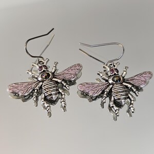 May include: A pair of silver-toned bee earrings. Each earring features a detailed bee design with purple-pink wings and a small, round, purple gem. The earrings have a hook closure.