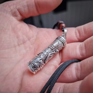 Puede incluir: Collar con colgante de plata con un diseño detallado y ornamentado que presenta una figura en posición sentada. El colgante está unido a un cordón negro.
