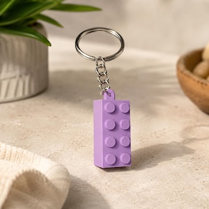May include: A lavender-colored rectangular building block keychain featuring a standard four-by-two stud pattern on the front surface. The plastic block is attached to a silver-toned metal split ring by a short, flexible metal chain link.