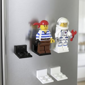 Puede incluir: Una colección de minifiguras estilo Lego, incluyendo un pirata y un astronauta, exhibidas en un refrigerador. El pirata tiene un pañuelo rojo y una camisa a rayas. El astronauta lleva un traje espacial y un casco blancos. También se muestran placas base compatibles con Lego en blanco y negro.
