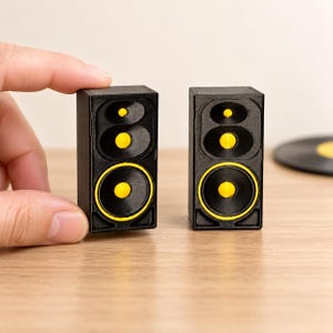 May include: Two miniature black speakers with bright yellow circular accents on the speaker cones. Each rectangular unit features a top tweeter and a larger bottom woofer, both with yellow centres, designed as small-scale replicas of audio equipment.