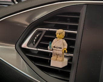 May include: A Lego figurine of a character from Star Wars, holding a blue lightsaber, is mounted on a car air vent. The figurine is wearing a white outfit with a tan belt and boots. The car's interior features a black dashboard and silver trim.