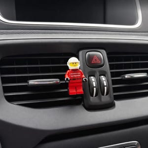 Könnte beinhalten: Eine rot-weiße Lego-Figur mit Helm ist an einer Lüftungsöffnung eines Autos befestigt. Die Figur befindet sich vor der Warnblinkertaste. Der Innenraum des Autos ist schwarz und grau.