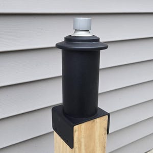 May include: A black metal post cap with a gray top, mounted on a wooden post. The post cap is designed to protect the top of the post from the elements.