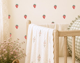 May include: A light-colored wall featuring a repeating pattern of small red strawberry decals. A wooden crib is positioned in front of the wall, draped with a cream-colored knit blanket featuring small embroidered pink floral sprigs.