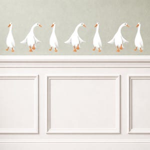 May include: A row of white goose decals with orange beaks and feet are shown against a pale green backdrop. The geese are in different poses, some with wings outstretched. The decals are placed above white wainscoting.
