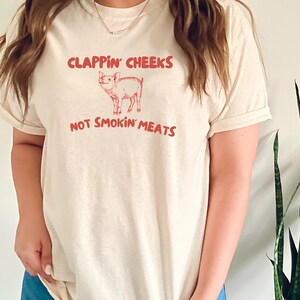 May include: A white t-shirt with a pink pig illustration and the text "Clappin' Cheeks Not Smokin' Meats".