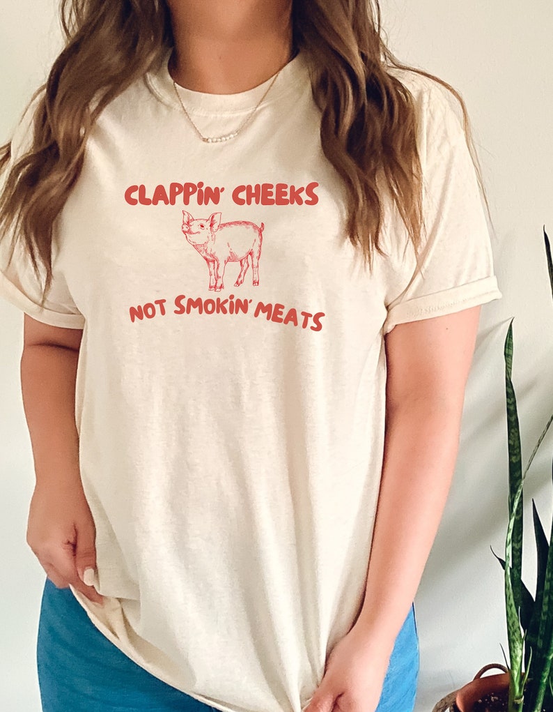 May include: A white t-shirt with a pink pig illustration and the text "Clappin' Cheeks Not Smokin' Meats".