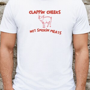 May include: White t-shirt with a red graphic of a pig and the text "CLAPPIN' CHEEKS NOT SMOKIN' MEATS".