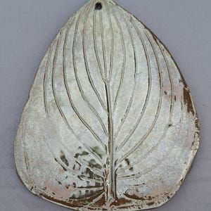 May include: A ceramic leaf-shaped wall hanging with a hole at the top for hanging. The leaf is a light, off-white color with a brown rim and detailed veining. The design is a stylized representation of a leaf.