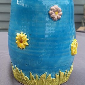 Puede incluir: Jarrón de cerámica turquesa hecho a mano, adornado con flores amarillas y rosas en relieve. Una banda de hierba verde adorna la base. Impresiones de hojas visibles. Medidas en centímetros.