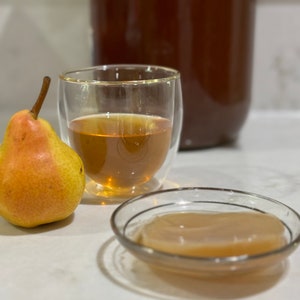 May include: A glass of kombucha with a pear and a kombucha scoby in a glass dish on a white countertop.