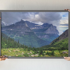 May include: A framed photograph of a mountain range with a cloudy sky. The mountains are green and rocky, with a valley in the foreground. The sky is a light blue with white clouds.