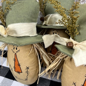 May include: Three stuffed fabric scarecrows with tan bodies, orange triangular noses, and stitched facial features. Each wears a sage green top hat adorned with a cream-colored fabric ribbon, a small brown jingle bell, and dried golden wheat sprigs.