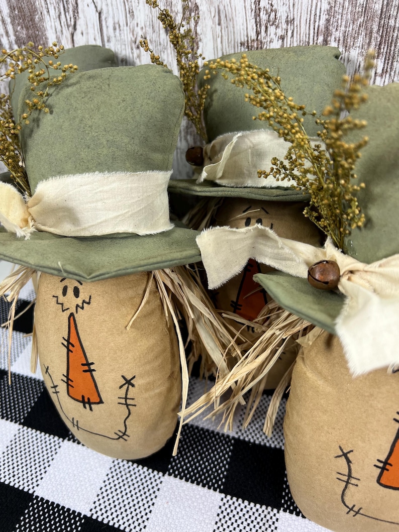 May include: Three stuffed fabric scarecrows with tan bodies, orange triangular noses, and stitched facial features. Each wears a sage green top hat adorned with a cream-colored fabric ribbon, a small brown jingle bell, and dried golden wheat sprigs.