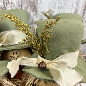 May include: A decorative figurine featuring a sage green fabric top hat adorned with a cream-colored frayed ribbon, a small rusted metal bell, and a sprig of dried yellow flowers. The base is made of tan material with a painted orange carrot shape.