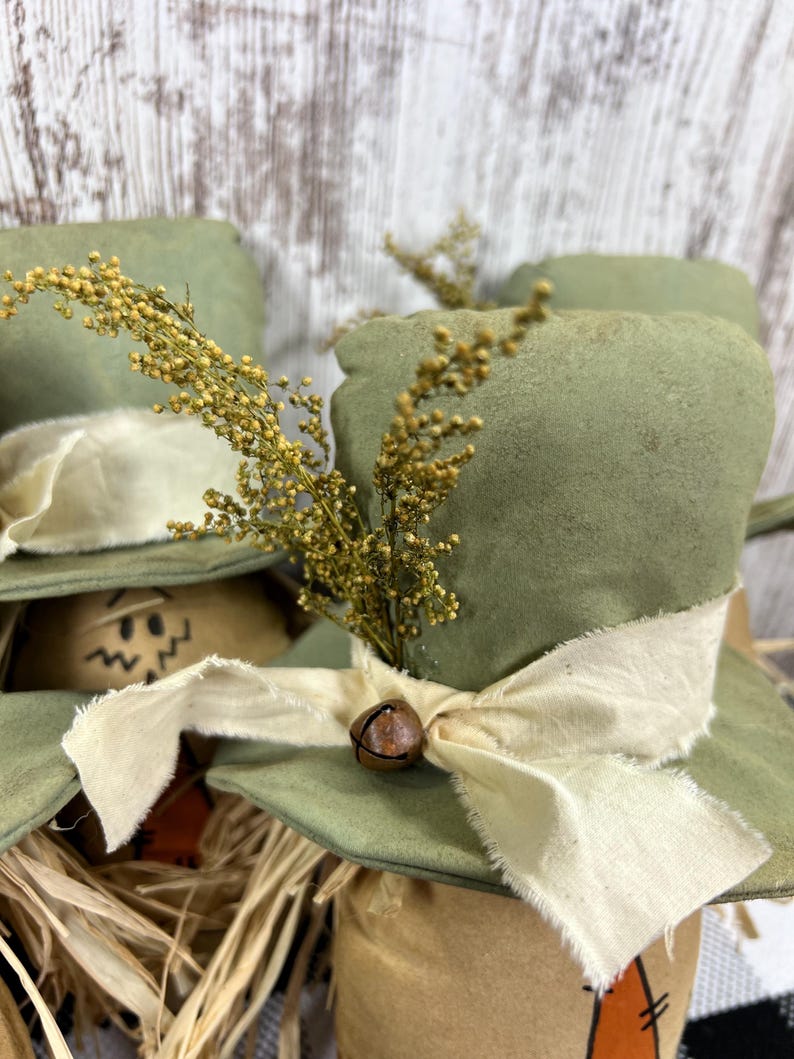 May include: A decorative figurine featuring a sage green fabric top hat adorned with a cream-colored frayed ribbon, a small rusted metal bell, and a sprig of dried yellow flowers. The base is made of tan material with a painted orange carrot shape.