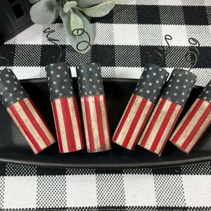 May include: Six cylindrical decorative items with a distressed American flag design, featuring red and white stripes and a blue field of stars. They are arranged on a black rectangular tray, set against a black and white checkered background.