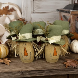 May include: A group of fabric scarecrow figures with tan bodies, stitched facial features, and orange triangular noses. Each figure wears a green fabric top hat adorned with a cream-colored ribbon and dried foliage accents, featuring straw hair.
