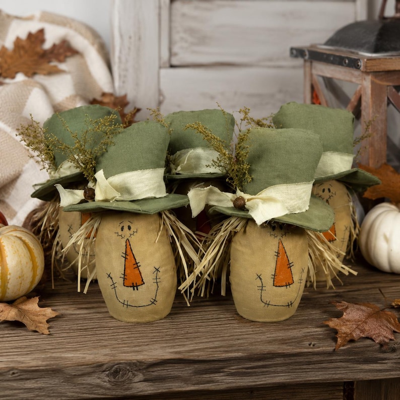 May include: A group of fabric scarecrow figures with tan bodies, stitched facial features, and orange triangular noses. Each figure wears a green fabric top hat adorned with a cream-colored ribbon and dried foliage accents, featuring straw hair.