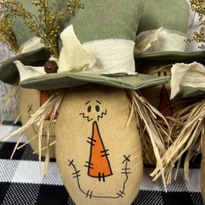 May include: A set of fabric scarecrow figures, each featuring a tan oval body with a hand-drawn face, a stitched orange carrot nose, and raffia hair. Each figure wears a green fabric top hat adorned with a cream-colored fabric ribbon and dried yellow floral sprigs.