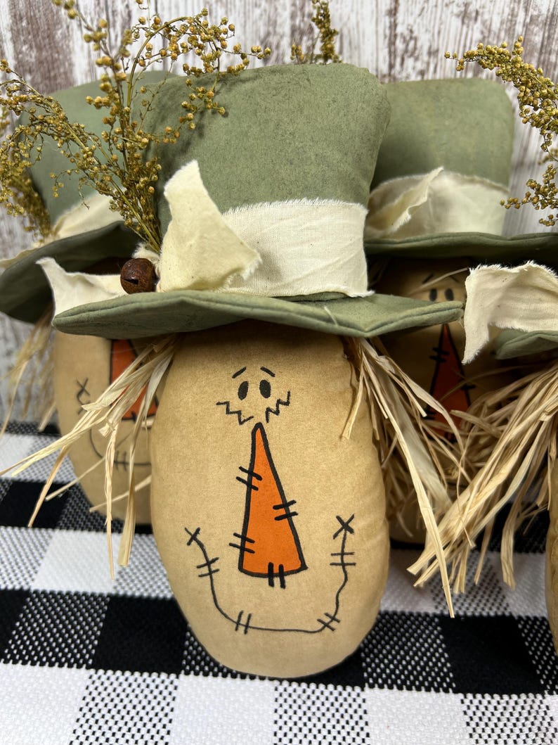 May include: A set of fabric scarecrow figures, each featuring a tan oval body with a hand-drawn face, a stitched orange carrot nose, and raffia hair. Each figure wears a green fabric top hat adorned with a cream-colored fabric ribbon and dried yellow floral sprigs.