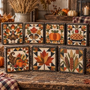 May include: A set of seven square wooden blocks featuring colorful autumn-themed quilt patterns. The designs include a turkey, acorns, a pumpkin, a pie, a cornucopia, a maple leaf, and corn stalks, all rendered in warm seasonal tones.